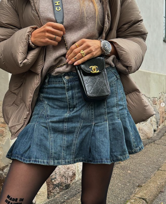 Medium blå denim nederdel med høj talje og flared design, båret med en brun jakke og sort taske.